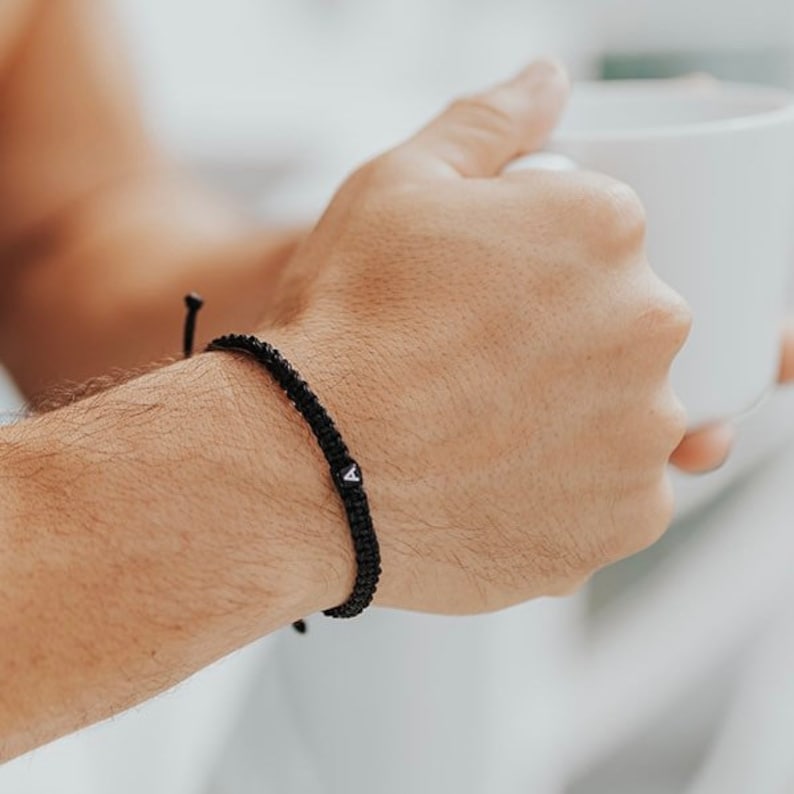 May include: Black braided bracelet with a small white charm featuring a black letter "A". The bracelet is worn on a wrist. The background is blurred, suggesting a casual setting.