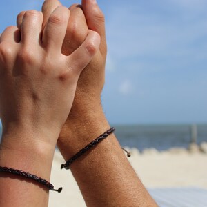 K&ouml;nnte beinhalten: Zwei Personen tragen passende braune geflochtene Armb&auml;nder an ihren Handgelenken. Die Armb&auml;nder sind mit einem Knoten gebunden und haben eine kleine Metallperle am Ende.