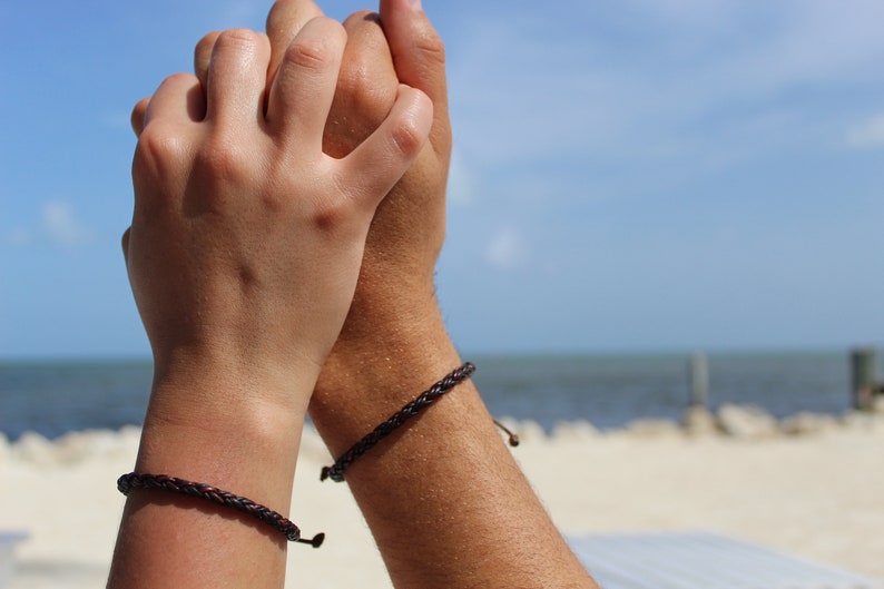 K&ouml;nnte beinhalten: Zwei Personen tragen passende braune geflochtene Armb&auml;nder an ihren Handgelenken. Die Armb&auml;nder sind mit einem Knoten gebunden und haben eine kleine Metallperle am Ende.