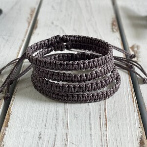 May include: A stack of five handmade macrame bracelets in a muted gray-brown color. Each bracelet is made of knotted cord and has adjustable ties. The bracelets are displayed on a white wooden surface.