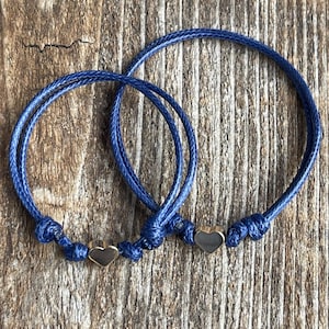 May include: Two blue friendship bracelets with gold heart charms. The bracelets are tied together with a knot.
