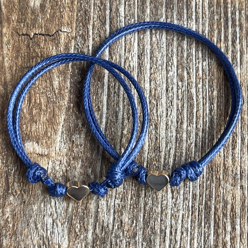 May include: Two blue friendship bracelets with gold heart charms. The bracelets are tied together with a knot.