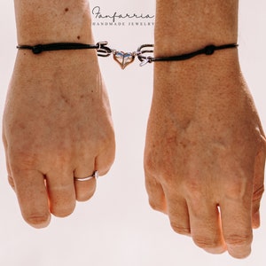 May include: Two wrists wearing matching black cord bracelets with a silver heart charm connecting them. The text "Fanfarria Handmade Jewelry" is visible above the bracelets.