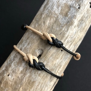 May include: Two bracelets, one black and one beige, are tied together in a knot. The bracelets are made of cord and are displayed on a piece of driftwood.