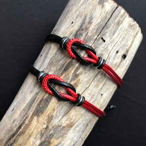 May include: Two matching bracelets with red and black rope-like cords. The bracelets are knotted in the center and have silver-colored metal accents. The bracelets are displayed on a piece of weathered wood.