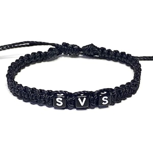 May include: Black braided bracelet with three black and white lettered beads spelling "SVS". The bracelet is made of a thick, woven material and is designed to be worn around the wrist.