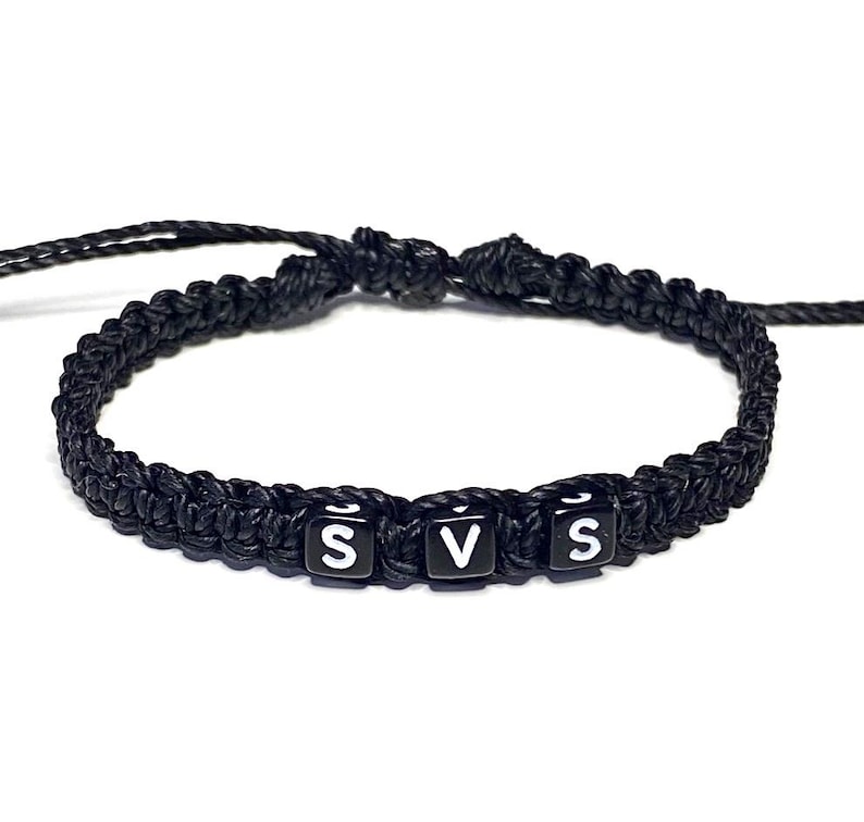 May include: Black braided bracelet with three black and white lettered beads spelling "SVS". The bracelet is made of a thick, woven material and is designed to be worn around the wrist.