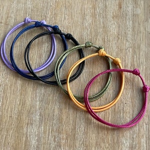 May include: Six colorful string bracelets in purple, black, dark green, orange, and pink. The bracelets are tied in a knot at the ends.