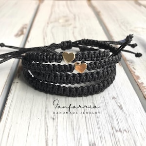 May include: A set of three black braided cord bracelets featuring small gold-toned heart charms. The bracelets have adjustable tie closures with loose ends. The text Fanfarria HANDMADE JEWELRY is printed in black below the bracelets.