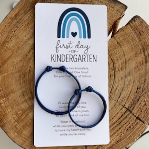 May include: Two navy blue cord bracelets with a knot closure are displayed on a white card. The card has a blue rainbow graphic and text that reads "first day of kindergarten." The card also includes a poem about the bracelets.