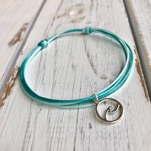 May include: A turquoise and white braided bracelet with a silver charm featuring a wave design.