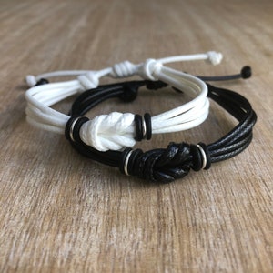 May include: Two friendship bracelets, one black and one white, with silver metal accents. The bracelets are tied in a knot.