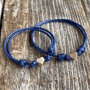 May include: Two blue friendship bracelets with gold heart charms. The bracelets are tied together and are on a wooden surface.