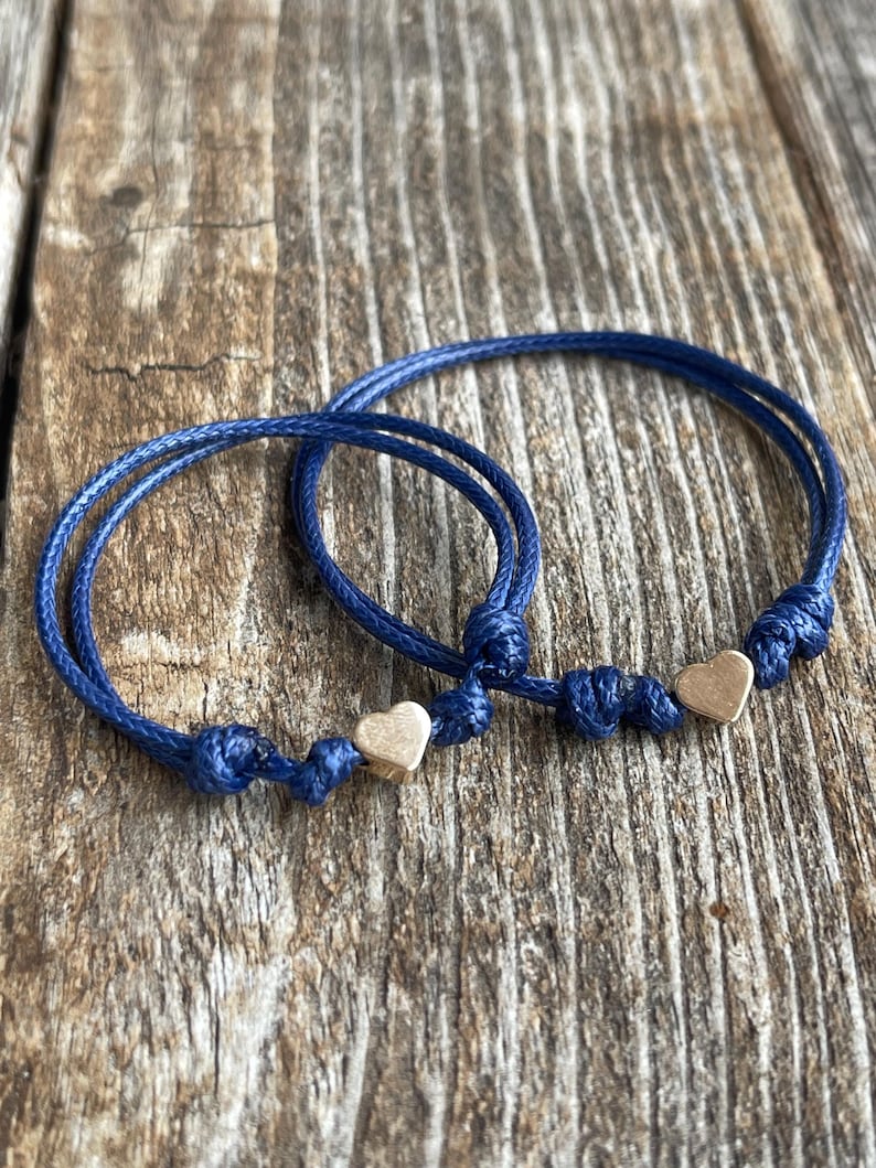 May include: Two blue friendship bracelets with gold heart charms. The bracelets are tied together and are on a wooden surface.