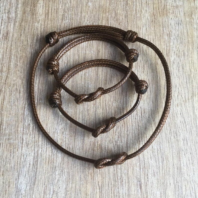 May include: Three brown cord bracelets with knotted ends. The bracelets are layered on top of each other.