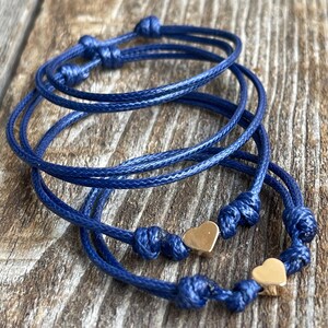 May include: Three blue cord bracelets with gold heart charms. The bracelets are knotted and layered on a wooden surface.