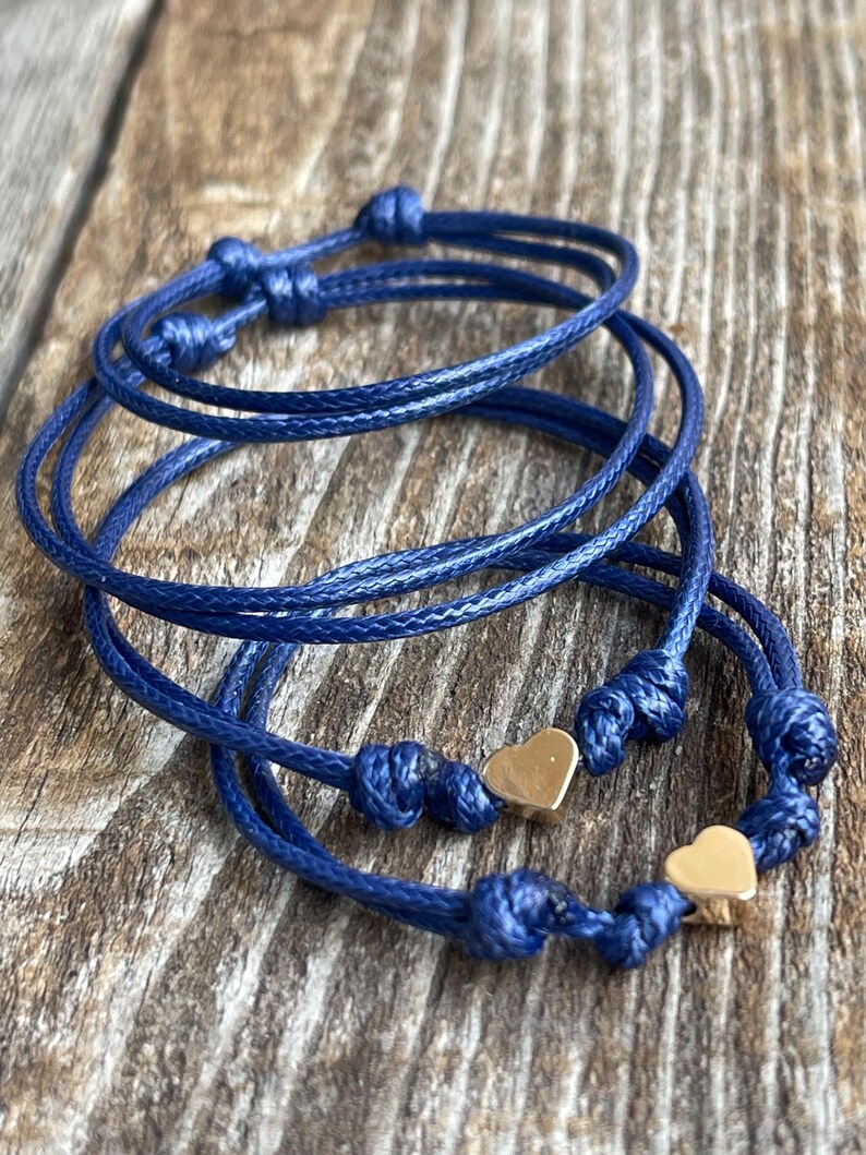 May include: Three blue cord bracelets with gold heart charms. The bracelets are knotted and layered on a wooden surface.