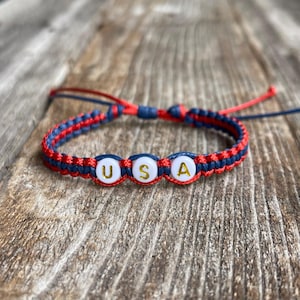 May include: Red and blue braided bracelet with the letters "USA" spelled out in gold lettering on white beads. The bracelet is on a wooden surface.