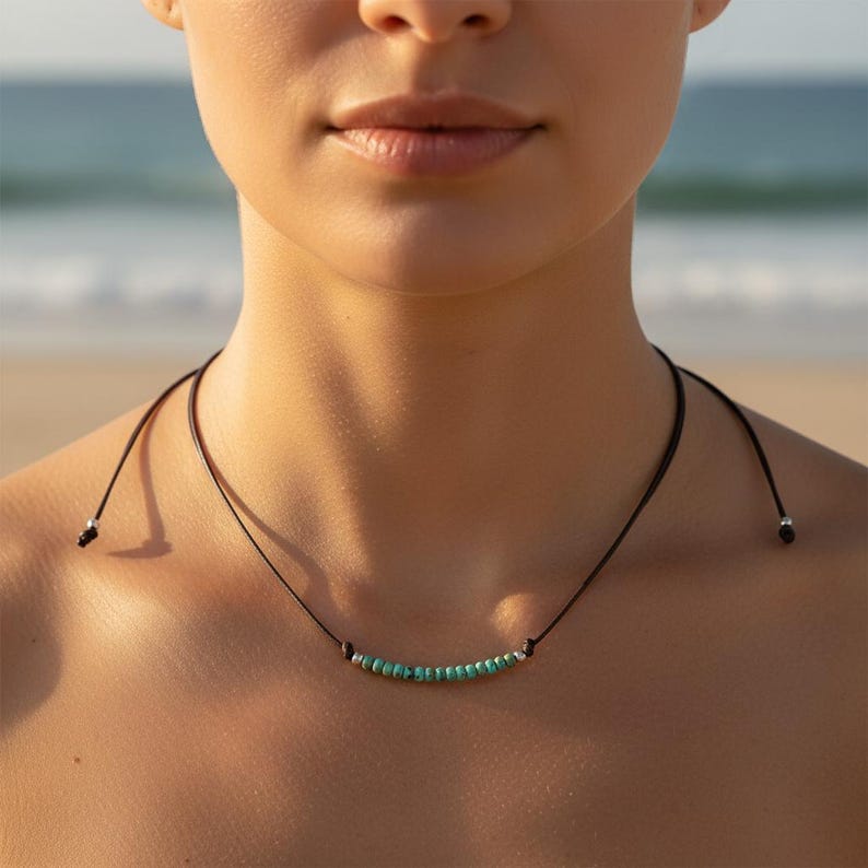 May include: A black cord necklace with turquoise beads and silver accents. The necklace is worn around the neck, with the beads resting on the collarbone. The background is a blurred beach scene.