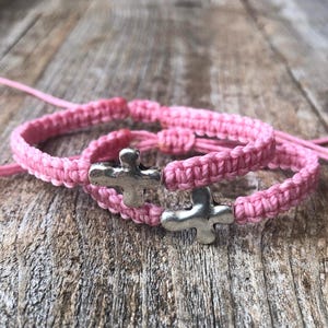 May include: Two pink braided bracelets with silver cross charms. The bracelets are tied together and are on a wooden surface.