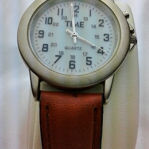 Könnte beinhalten: Eine silberfarbene Armbanduhr mit einem braunen Lederarmband. Das Zifferblatt der Uhr ist weiß mit schwarzen Zahlen und Zeigern. Auf dem Zifferblatt der Uhr steht auch das Wort "TIME".