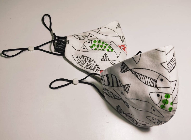 May include: Two white face masks with black elastic ear loops and a black and white fish pattern. One mask has green and red accents on the fish.