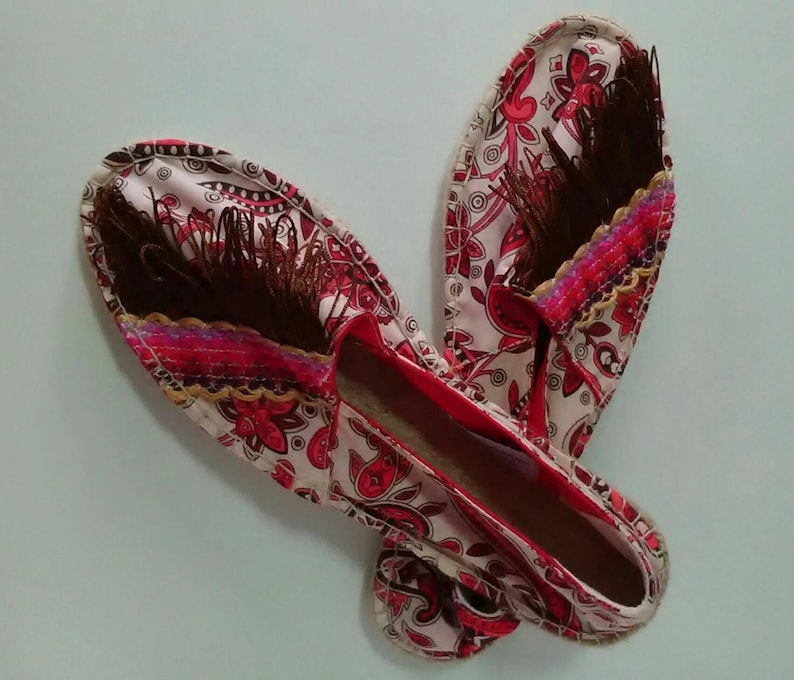 May include: A pair of red and white patterned espadrilles with brown tassels. The espadrilles have a red trim and a white sole.