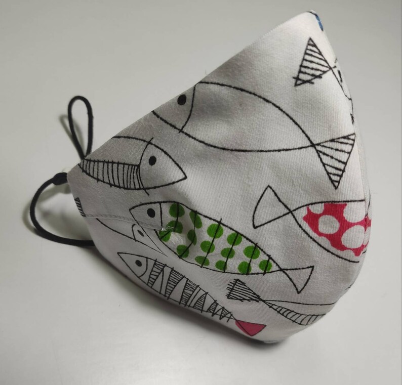 May include: White face mask with a black elastic band. The mask has a pattern of black and white fish with green and red accents.