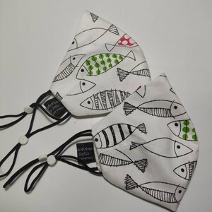 May include: Two white face masks with black elastic ear loops. Both masks feature a white background with black and white fish designs. One mask has green and red polka dots on the fish. The masks have the text "de You Sofia" printed on them.