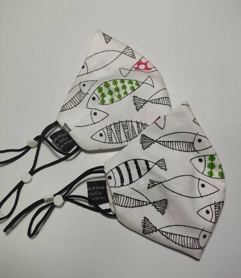 May include: Two white face masks with black elastic ear loops. Both masks feature a white background with black and white fish designs. One mask has green and red polka dots on the fish. The masks have the text "de You Sofia" printed on them.