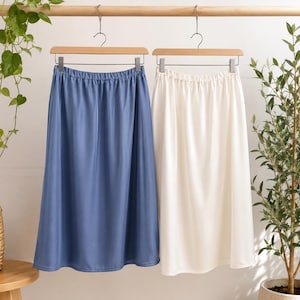 May include: Two midi skirts hanging on wooden hangers. One skirt is a solid dusty blue colour, and the other is a solid off-white colour. Both skirts feature an elasticated waistband and a smooth, flowing fabric that falls to a mid-calf length.