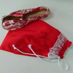 May include: A red fabric drawstring bag with white lace trim and white drawstrings. The bag is lying on a white surface with a floral patterned espadrille shoe next to it.