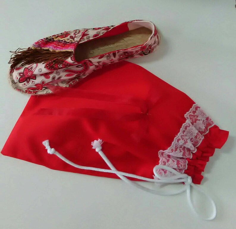 May include: A red fabric drawstring bag with white lace trim and white drawstrings. The bag is lying on a white surface with a floral patterned espadrille shoe next to it.