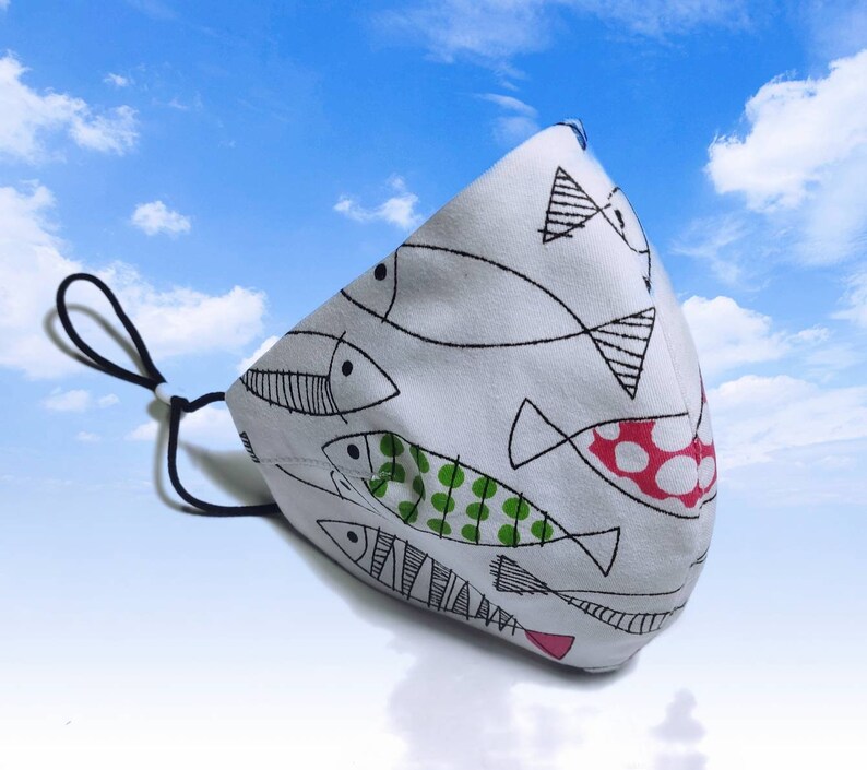 May include: White face mask with a black elastic band. The mask has a pattern of black and white fish with green and red accents.