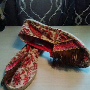 May include: A pair of red and white espadrille shoes with a paisley pattern and brown fringe. The shoes have a rope sole and a red trim.