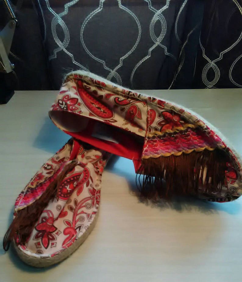 May include: A pair of red and white espadrille shoes with a paisley pattern and brown fringe. The shoes have a rope sole and a red trim.