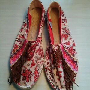 May include: A pair of red and white patterned espadrilles with brown tassels. The espadrilles have a white canvas upper with a red and white paisley pattern. The tassels are made of brown fabric and are attached to the front of the shoes.