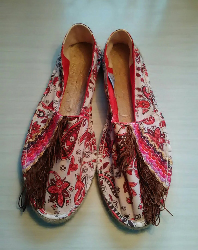 May include: A pair of red and white patterned espadrilles with brown tassels. The espadrilles have a white canvas upper with a red and white paisley pattern. The tassels are made of brown fabric and are attached to the front of the shoes.