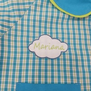 Pode incluir: Um tecido xadrez azul e branco com uma borda verde. O tecido tem um remendo em forma de nuvem branca com o nome "Mariana" bordado nele.