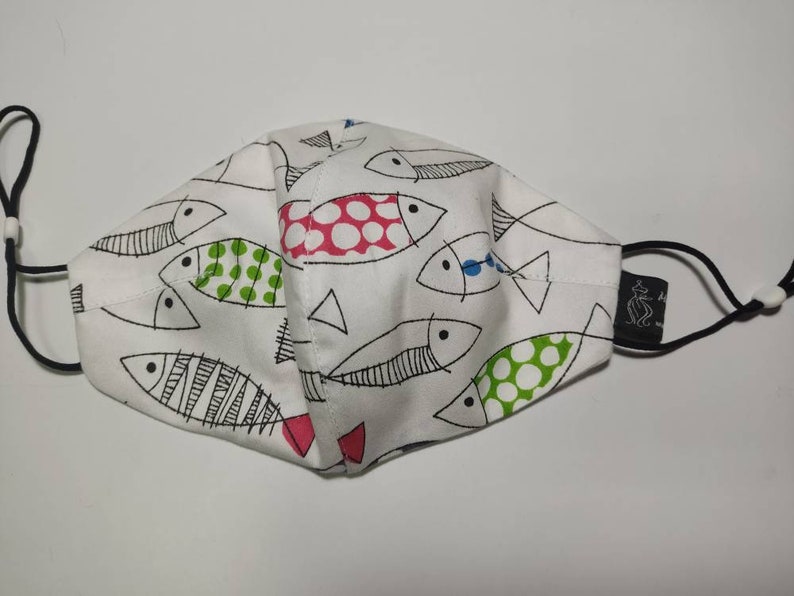 May include: White face mask with a black elastic band and a black label. The mask has a pattern of black and white fish with red, green, and blue accents.