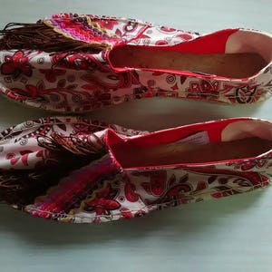 May include: A pair of white canvas espadrilles with a red and pink paisley pattern. The shoes have brown tassels and a red trim around the opening.