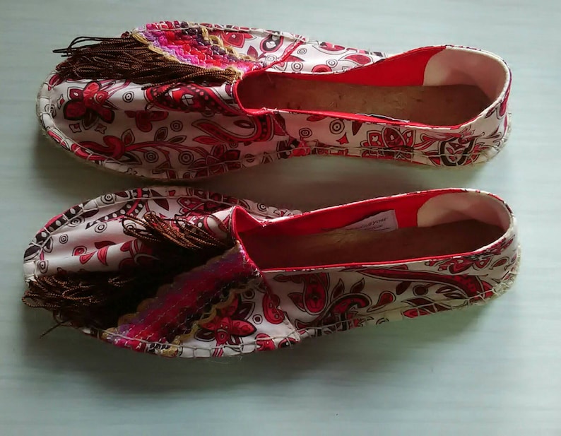 May include: A pair of white canvas espadrilles with a red and pink paisley pattern. The shoes have brown tassels and a red trim around the opening.