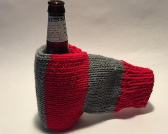 Può includere: Un copribottiglia di lana rosso e grigio con una bottiglia di birra all'interno. L'etichetta della bottiglia recita "Prism Ale".