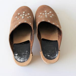 May include: A pair of brown leather clogs with white studs. The clogs have a black leather insole and a wooden base.