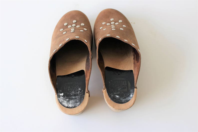 May include: A pair of brown leather clogs with white studs. The clogs have a black leather insole and a wooden base.