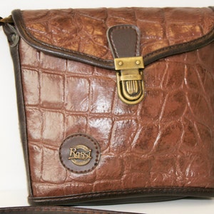 May include: A brown leather crossbody bag with a crocodile embossed pattern. The bag has a flap closure with a brass buckle and a small circular logo with the text "Rossi" on the front.