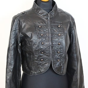 May include: A black faux leather jacket with a cropped hem and a military-inspired design. The jacket features a stand-up collar, a button-up front closure, and rows of decorative buttons down the front.