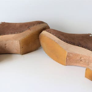 May include: A pair of brown suede clogs with wooden heels. The clogs have a stitched seam around the top edge and a small metal rivet on the side.