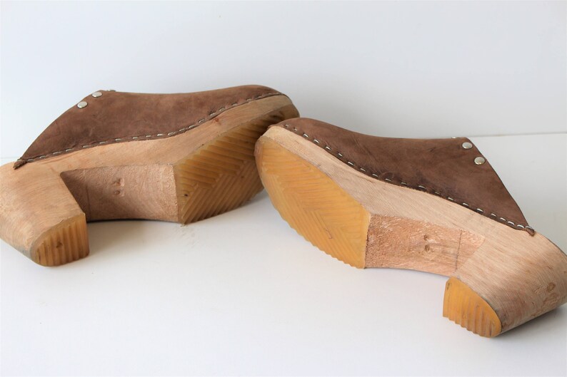 May include: A pair of brown suede clogs with wooden heels. The clogs have a stitched seam around the top edge and a small metal rivet on the side.
