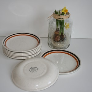 May include: A stack of six white ceramic plates with brown and orange trim. The plates are speckled with brown and white flecks. The plates are stacked on a white surface.